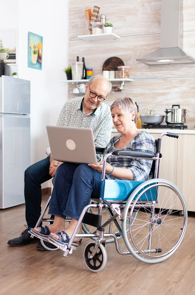 Senior couple at home looking at a laptop, with the man seated in a manual wheelchair to support independent living.