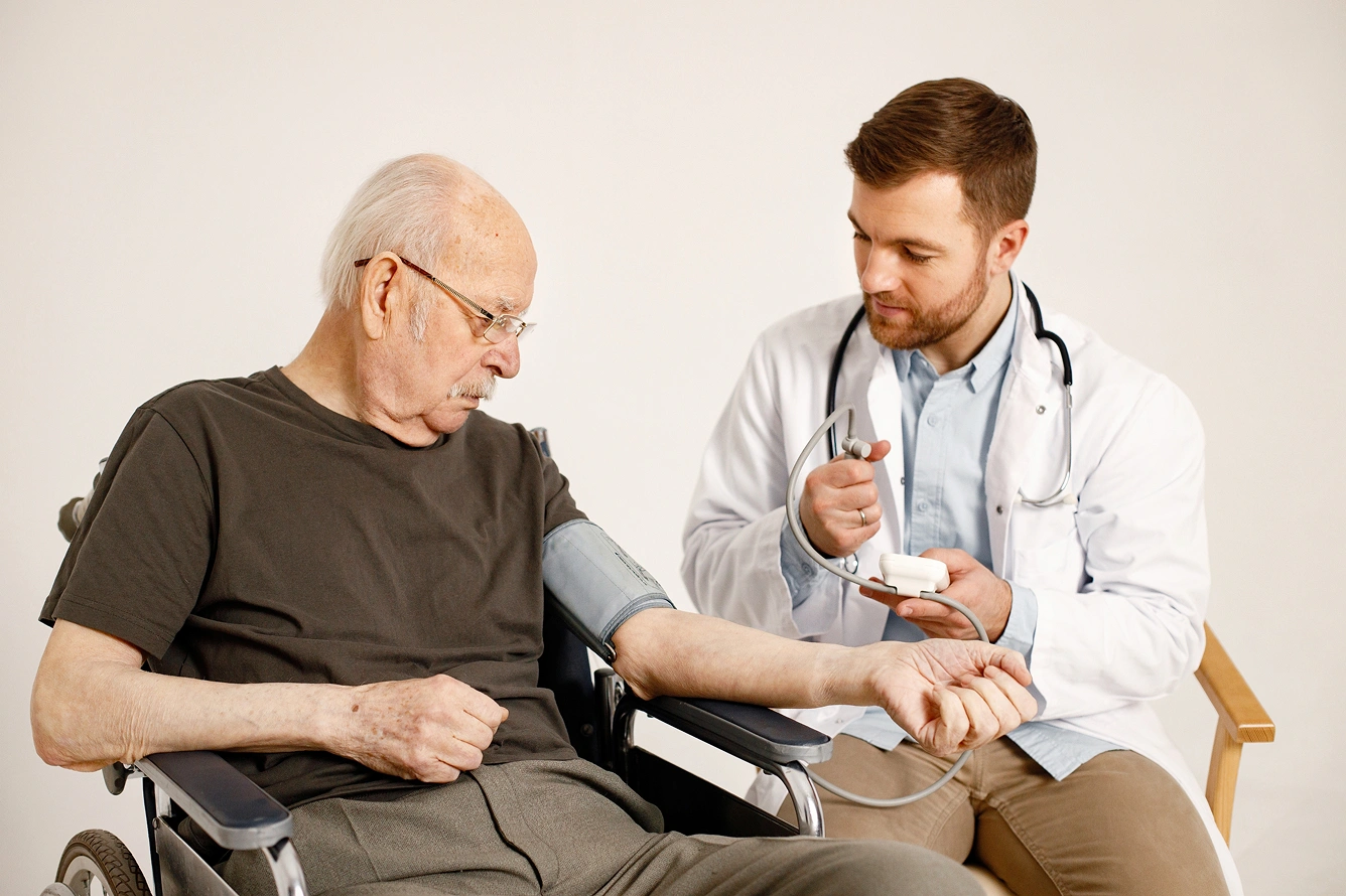 A doctor taking the blood pressure of an elderly male patient seated in a wheelchair, using professional diagnostic medical equipment.